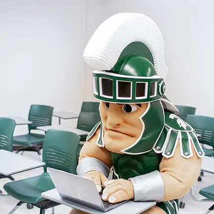 MSU mascot, Sparty, working on a laptop while seated in a classroom 